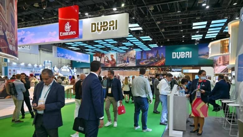 People walking and interacting at a trade show with stands displaying "Dubai" and "Emirates" signs, featuring various promotional materials and a bright ceiling display.
