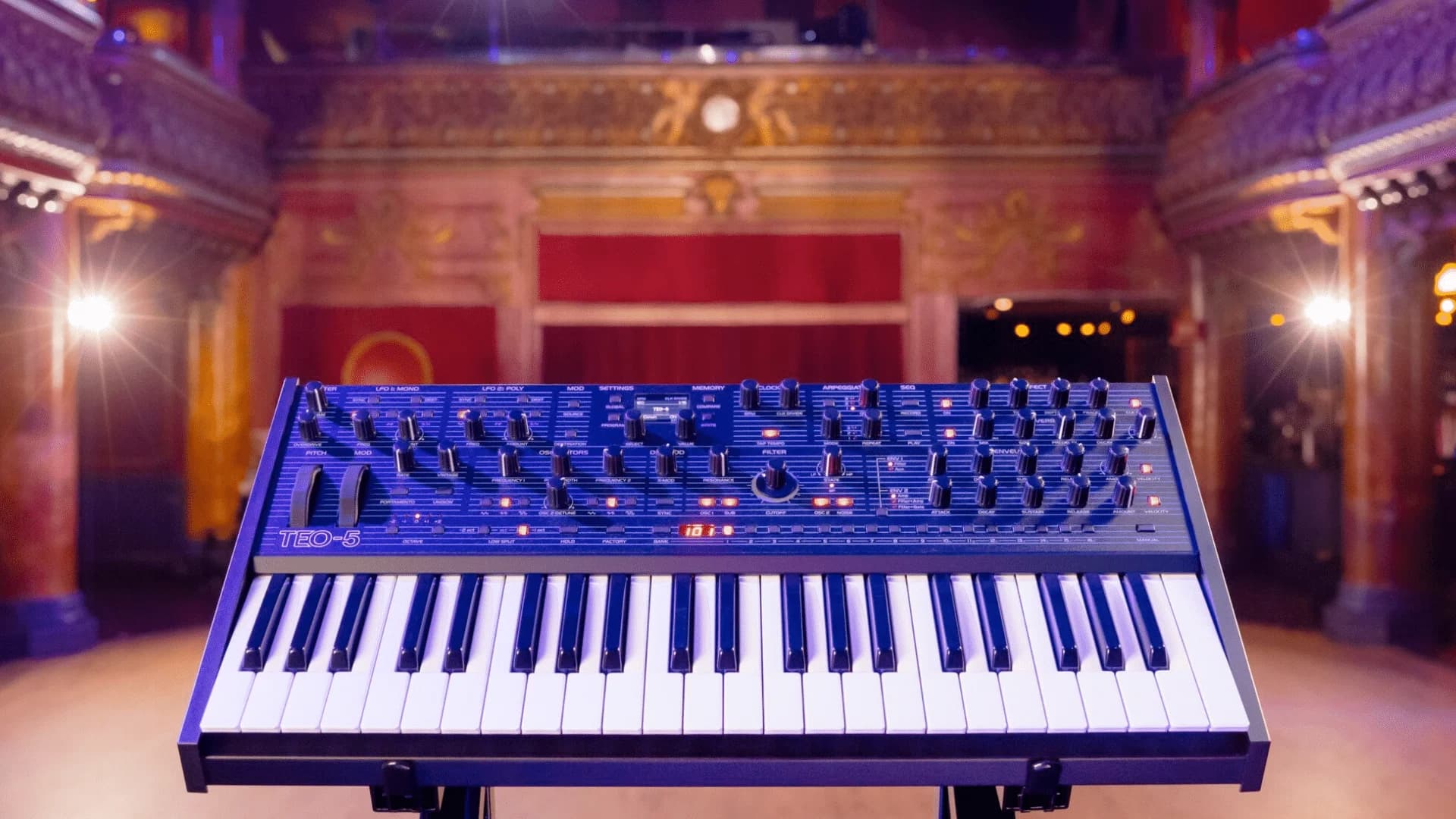 A synthesizer on a stand in a dimly lit theater with ornate details and a red velvet curtain in the background.