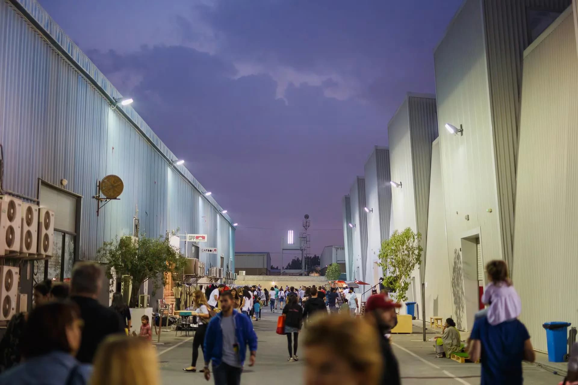 The image shows a busy outdoor scene in an industrial area at dusk. Tall warehouse-like buildings with corrugated metal walls line both sides of the walkway, illuminated by outdoor lights. A crowd of people, including a person carrying a child on their shoulders, are walking along the path, creating a bustling atmosphere under the twilight sky.