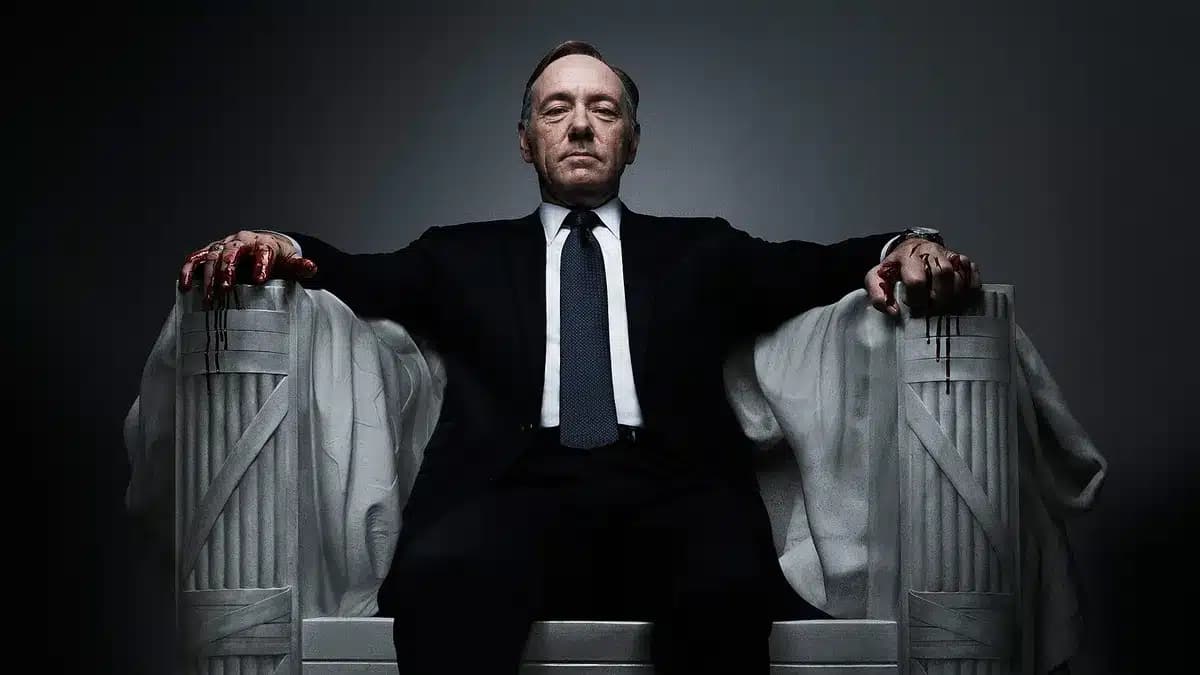 A man in a suit sits on a large chair with his arms resting on the armrests, hands stained with dripping blood.