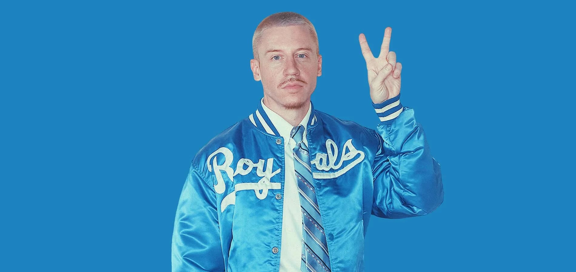 A person wearing a shiny blue jacket with the word 'Royals' printed in white, holding up a peace sign against a solid blue background.