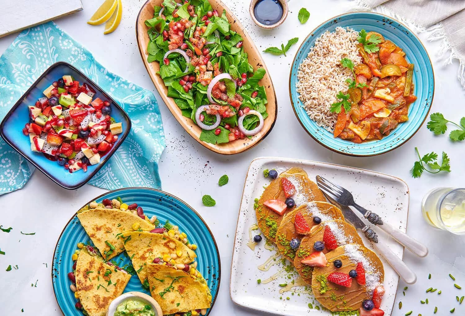 An array of vibrant dishes on a table, including a plate of pancakes with berries, a colorful salad with greens and onions, a bowl of rice with vegetable curry, a blue bowl with chopped fruits, and a plate of quesadillas with corn and beans, accompanied by a small dish of guacamole.
