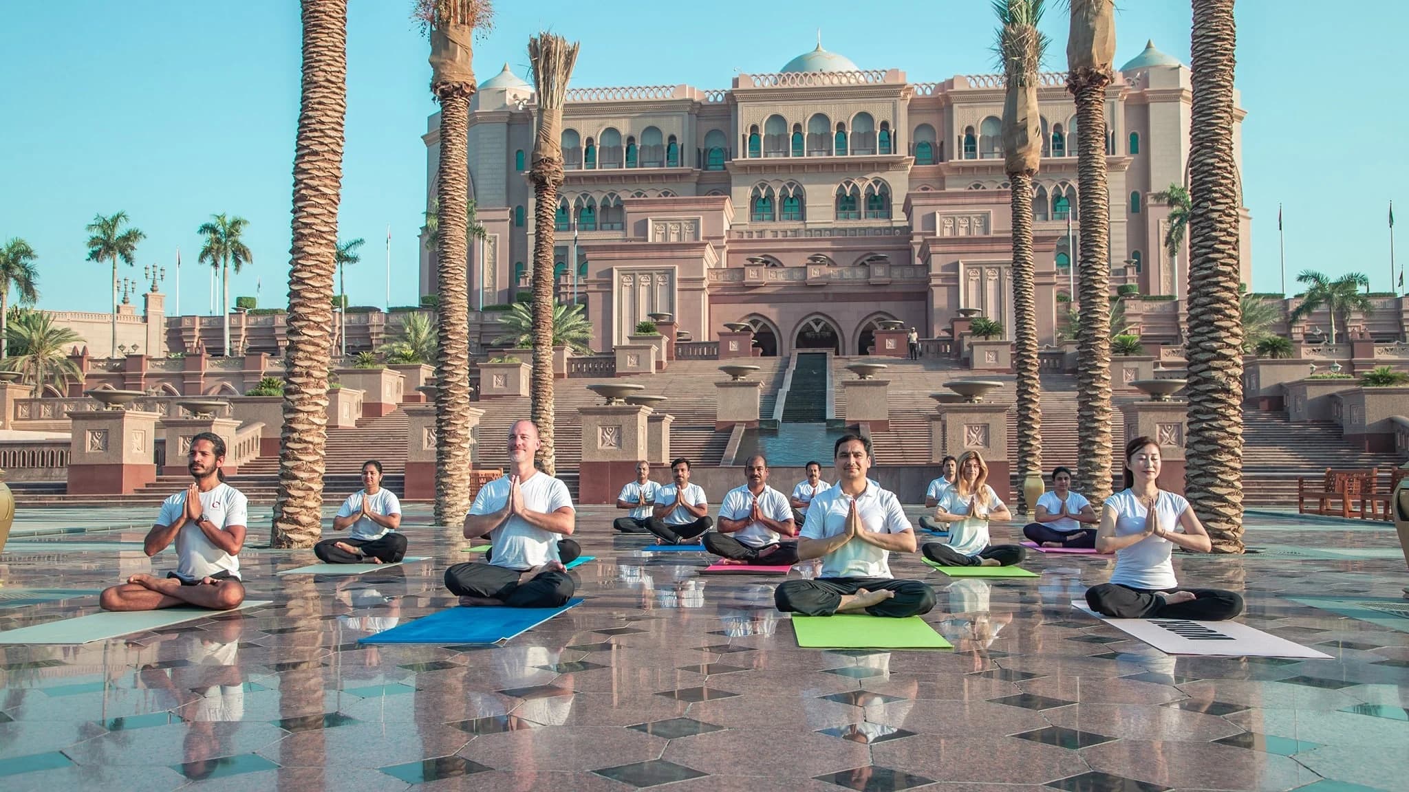 A group of people practicing yoga on colorful mats in front of an ornate building with palm trees, under a clear blue sky.