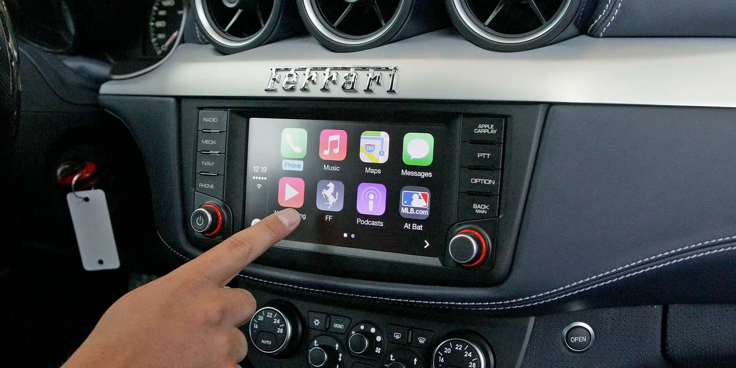 A hand touches the display of a car's infotainment system featuring various app icons, with the brand name 'Ferrari' above it.