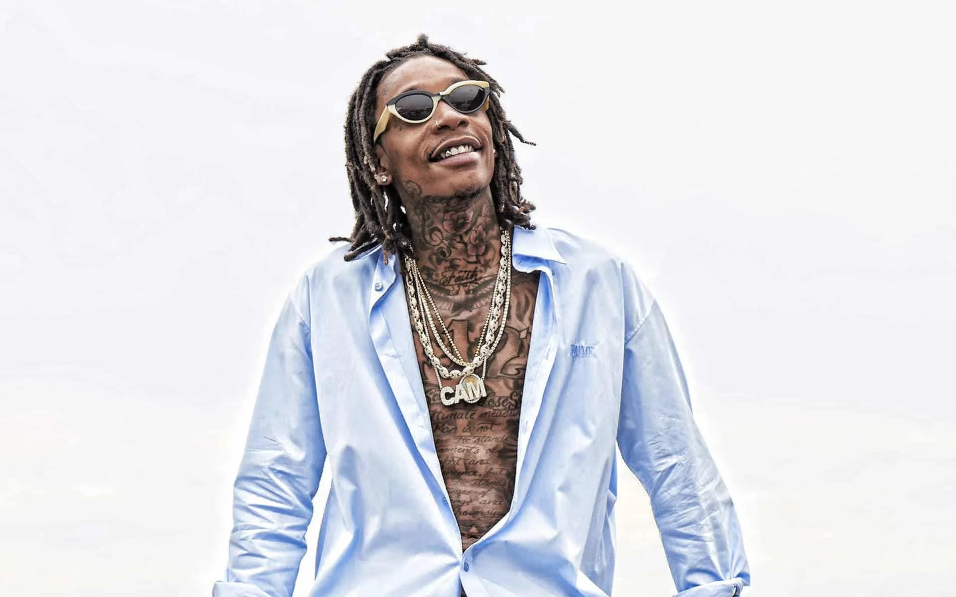 Man with dreadlocks wearing sunglasses, an open light blue shirt, and multiple necklaces, smiling and looking upward.