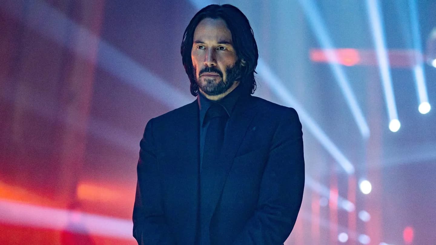 A man with long hair and a beard wearing a black suit stands against a background of colorful lights and beams.