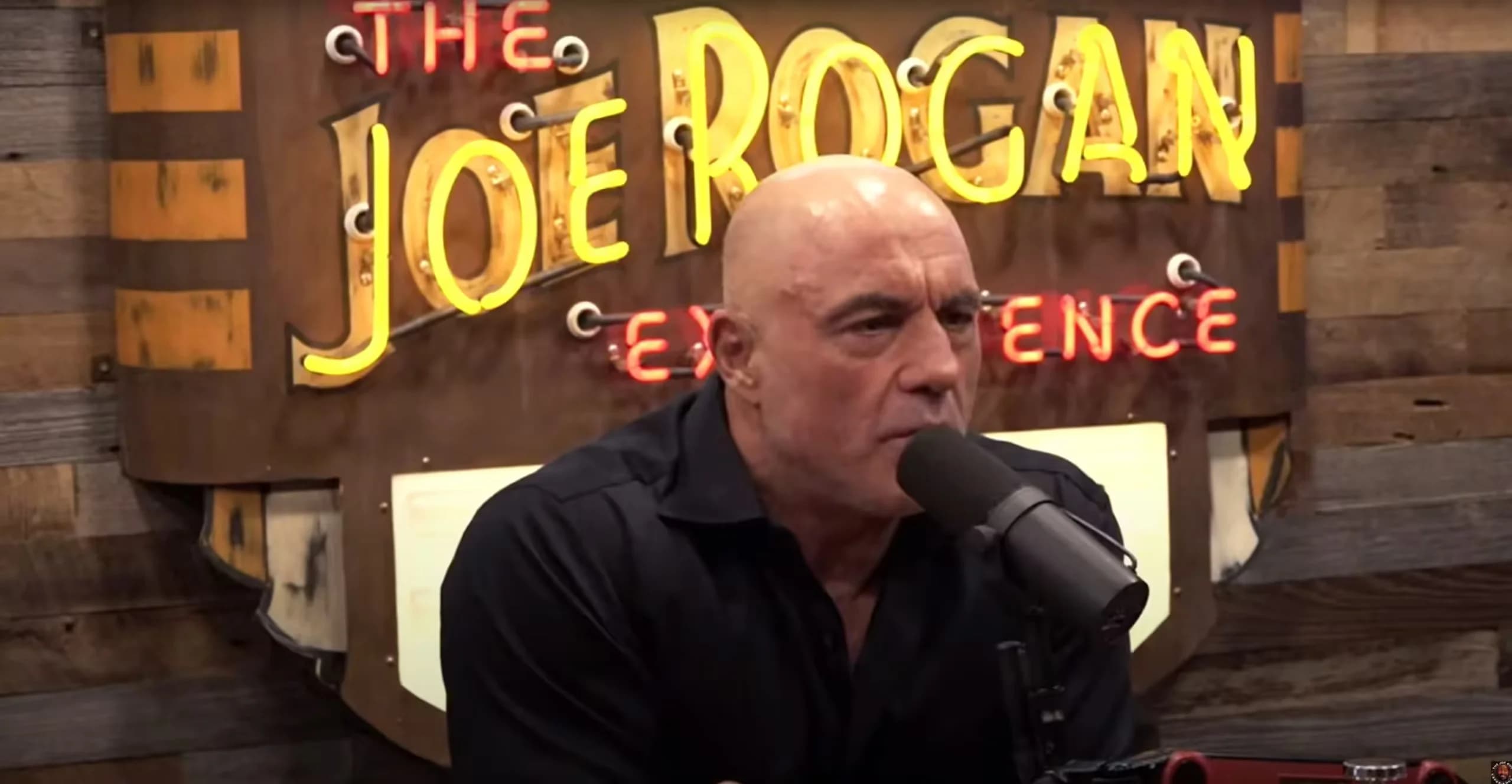 A man speaking into a microphone with a wooden wall and a neon sign behind him that says "The Joe Rogan Experience."