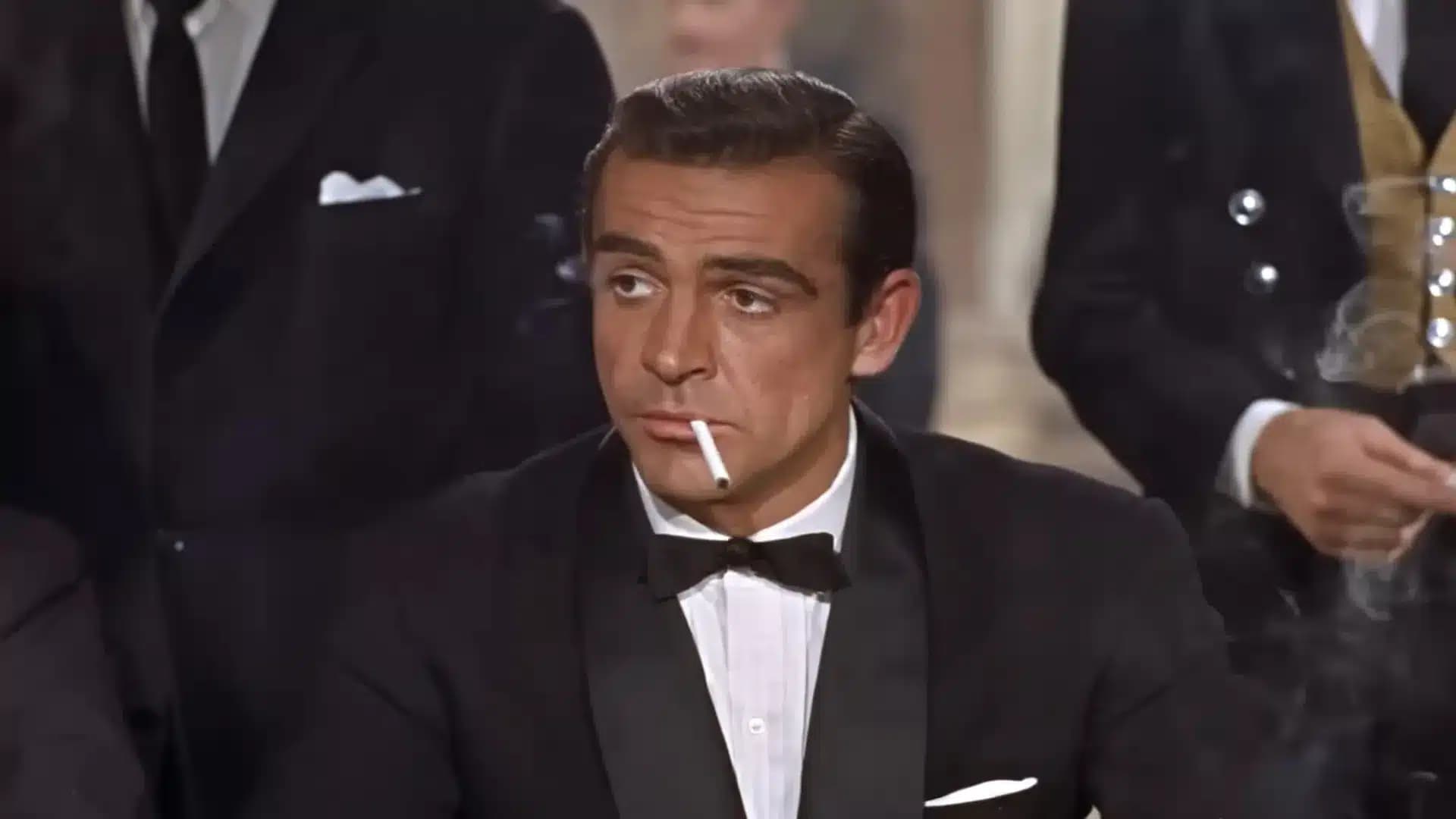 A man in a tuxedo with a bow tie is seated at a table, holding a cigarette in his mouth, surrounded by others in formal attire.
