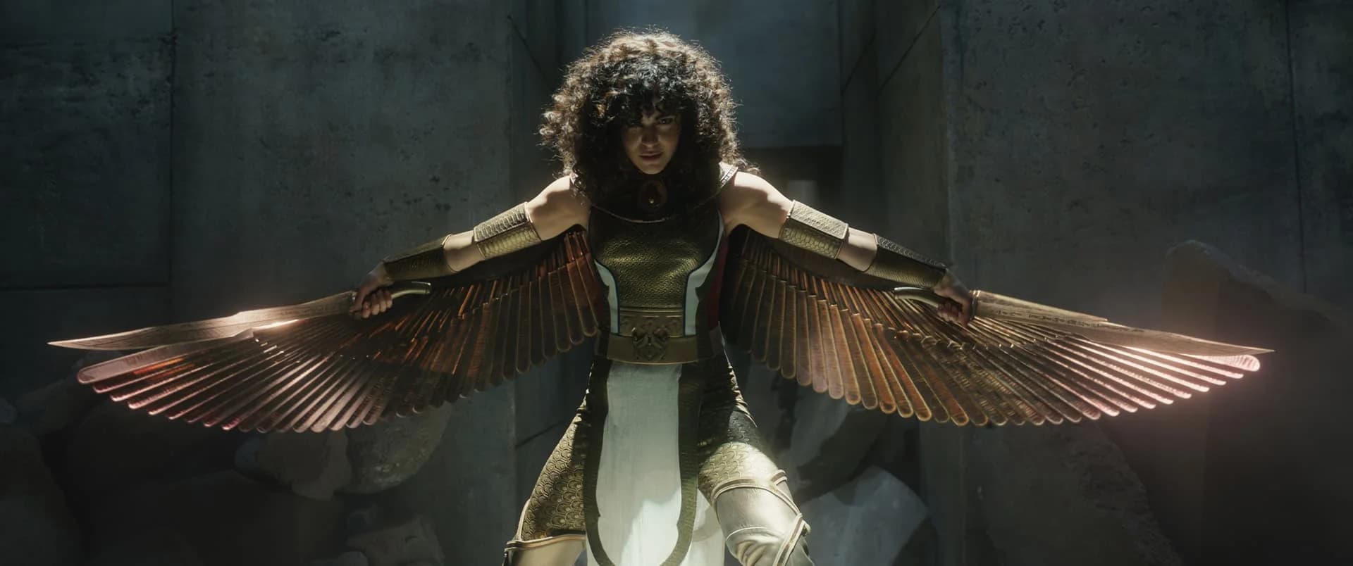 A person wearing an intricately designed costume with metallic wings extended, standing in a dramatic pose in a dimly lit, stone-walled environment.