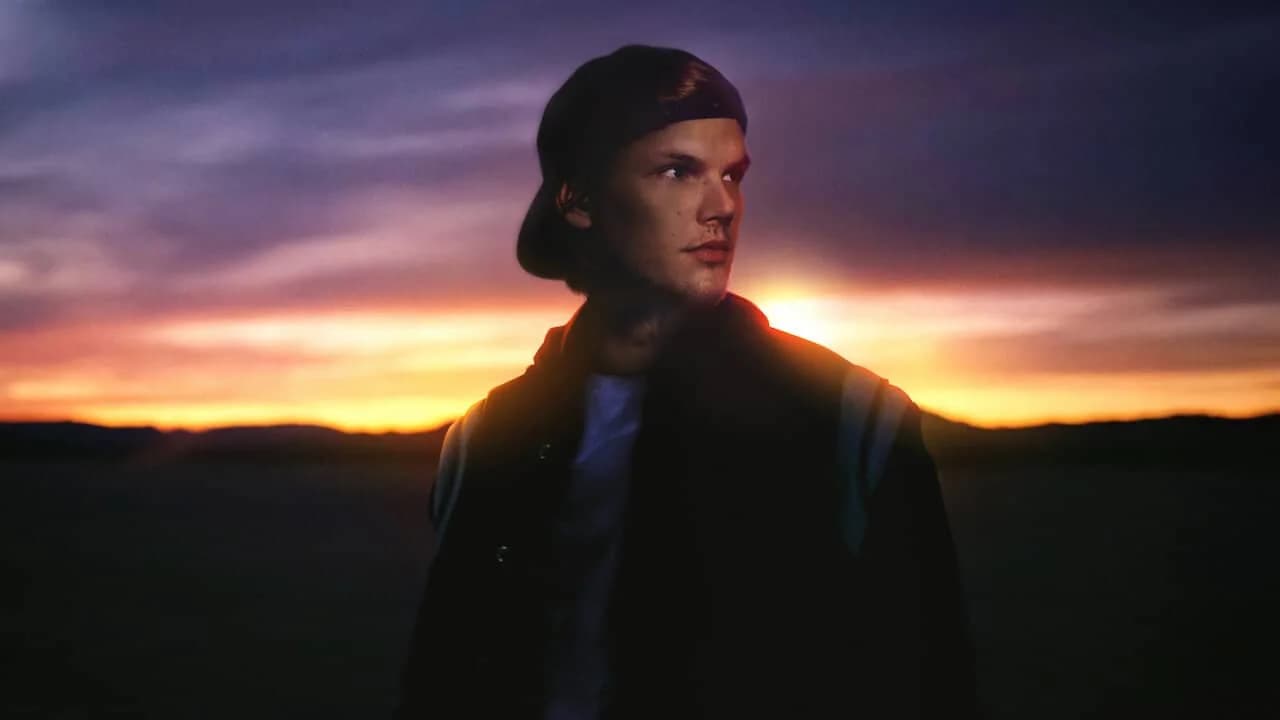 A person wearing a cap looks thoughtfully to the side, standing against a dramatic sunset with a gradient of orange and purple hues in the sky.