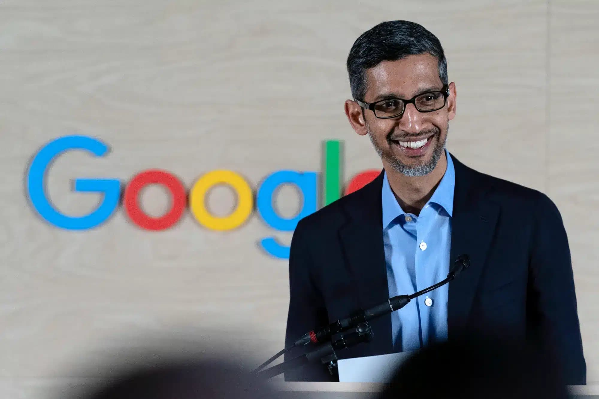 A person in a suit and glasses smiling while speaking at a podium, with a blurred Google logo in the background on a wooden panel.