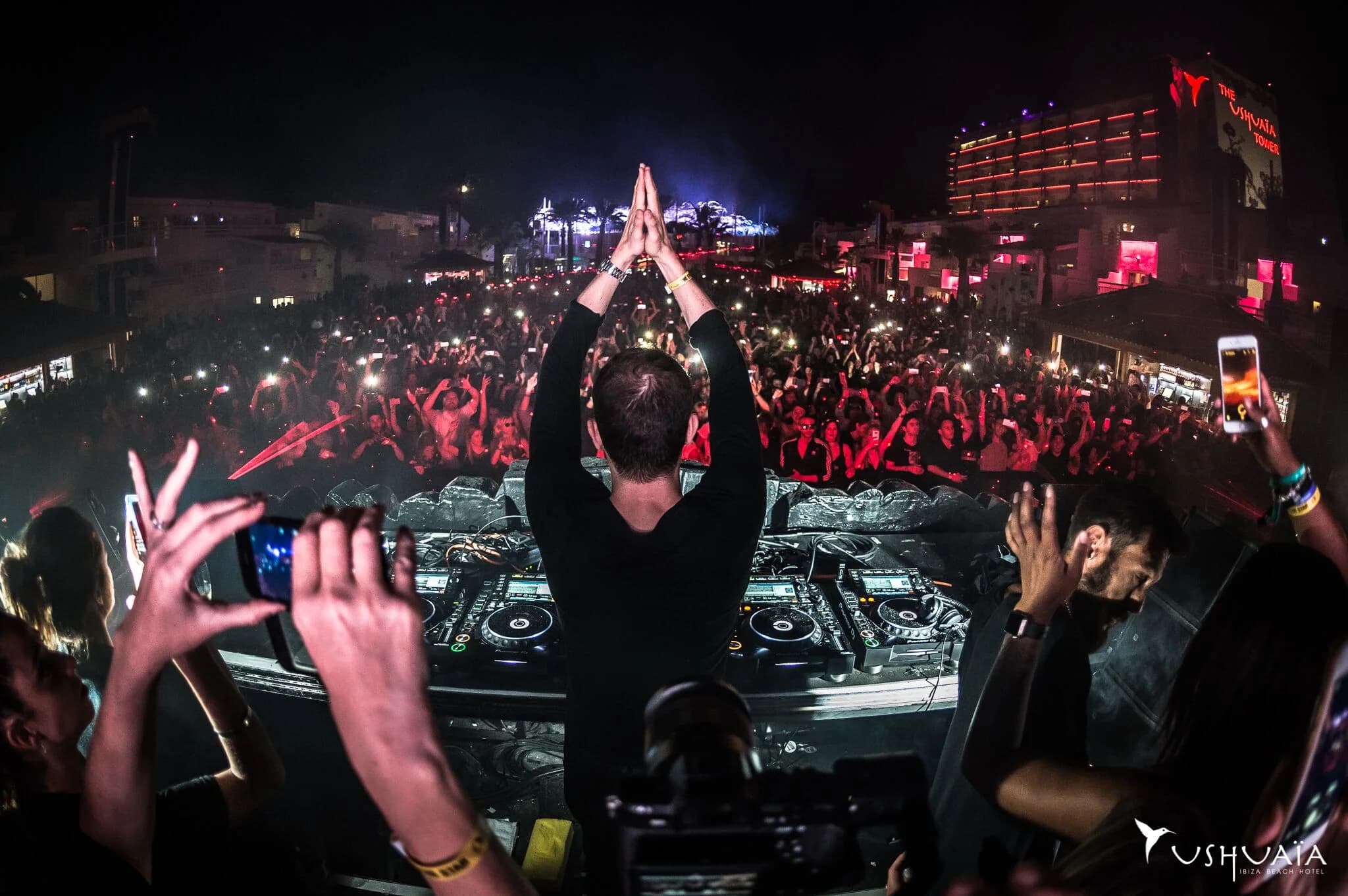 A DJ performs at an outdoor night event with a large crowd in front, illuminated by lights and camera flashes. The scene includes multiple DJ decks and visual effects, set against a backdrop of a brightly lit hotel building displaying the name 'Ushuaïa.' People hold up smartphones, capturing the moment.