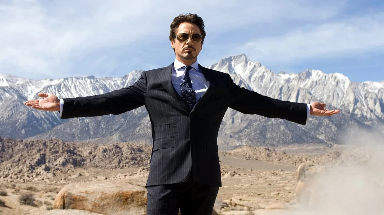 A man in a dark pinstripe suit, wearing sunglasses, stands with arms outstretched in front of a mountainous landscape with snow-capped peaks under a clear blue sky.