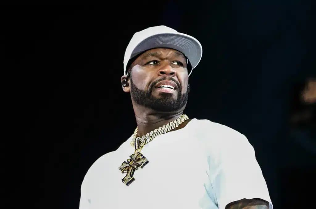 A man wearing a white cap and shirt, with a prominent beard and a large gold chain visible around his neck, looking slightly to the side with an expressive facial expression against a dark background.