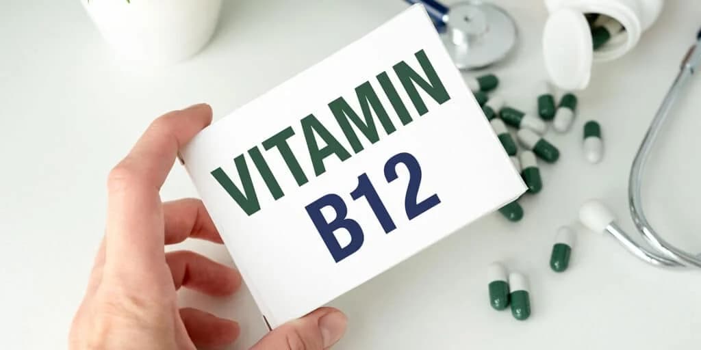 A hand holding a box labeled 'VITAMIN B12' with green text, surrounded by scattered green and white capsules and a stethoscope in the background.
