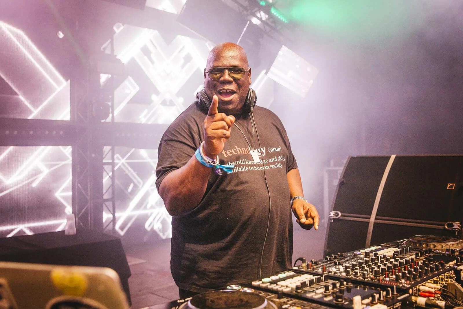 A DJ, wearing sunglasses and a black t-shirt with a definition of 'technology' printed on it, raises a finger while performing at a festival. He stands behind a DJ booth with various equipment and colorful stage lighting in a smoky atmosphere.
