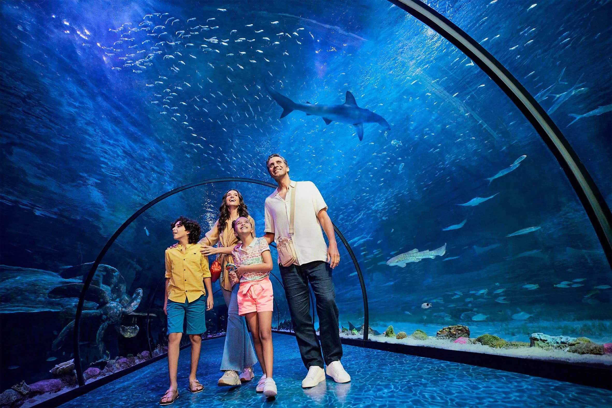 A family of four, including two children, is exploring an underwater aquarium tunnel with an array of fish swimming above them, including a shark. The adults are smiling, wearing casual clothing, while the children are holding colorful toys.