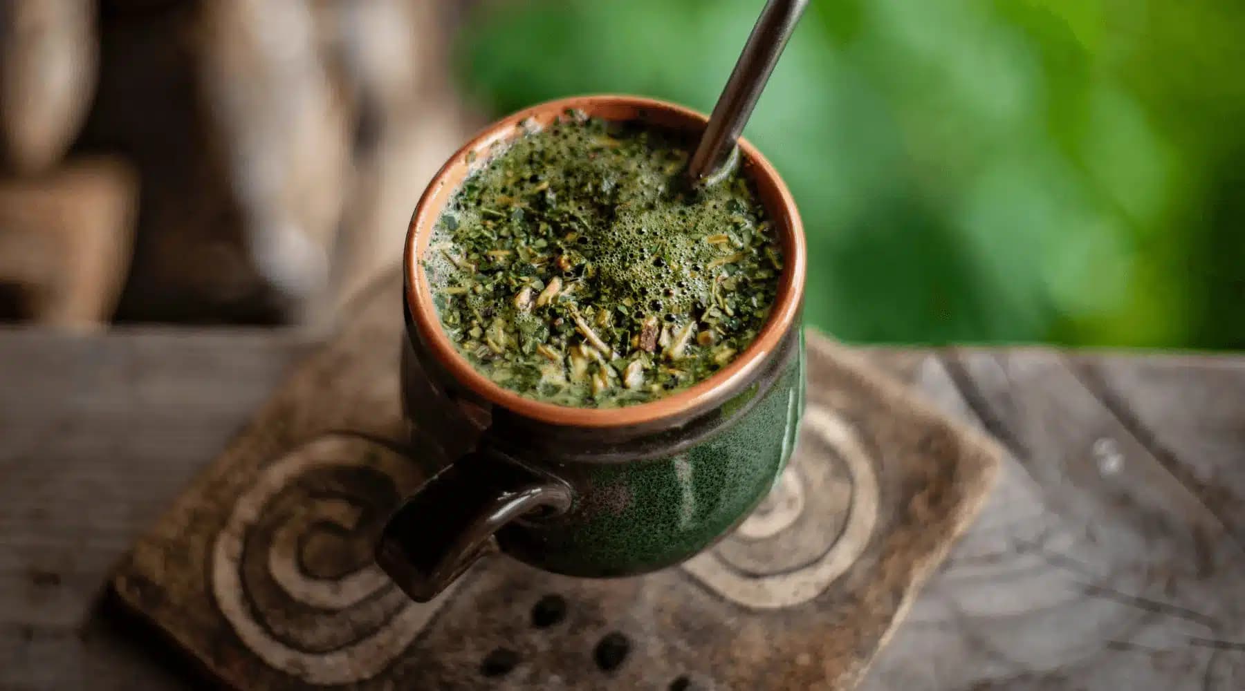 A close-up of a traditional ceramic mug filled with dark green, frothy beverage, resting on a wooden surface with intricate designs.
