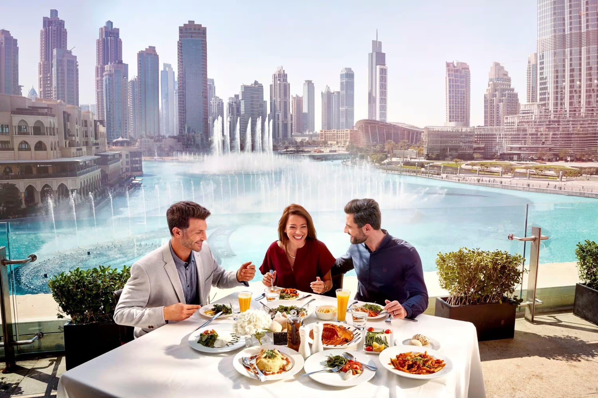 Three people enjoy a meal at an outdoor table overlooking a city skyline and large fountain.