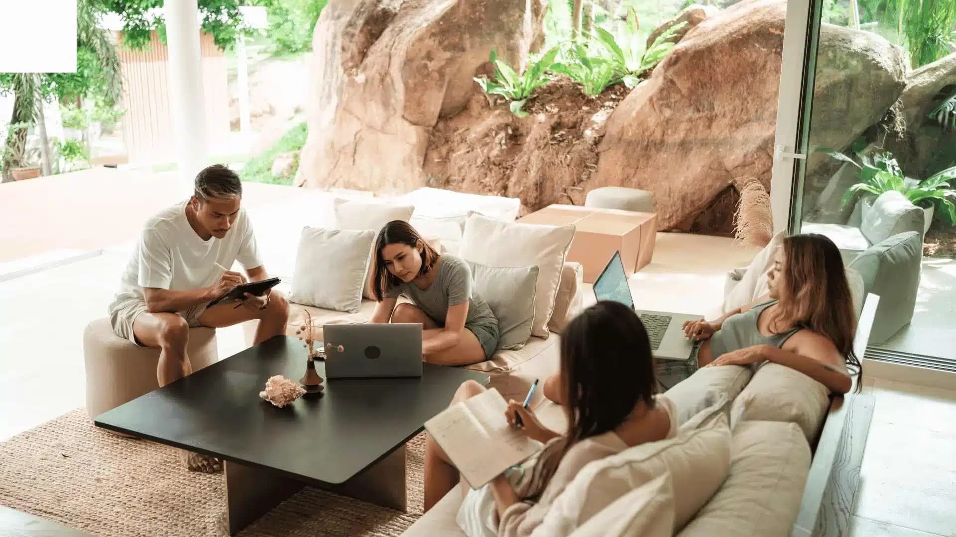 Four people sit in a modern living room with laptops and tablets, surrounded by large windows overlooking greenery and rocks.