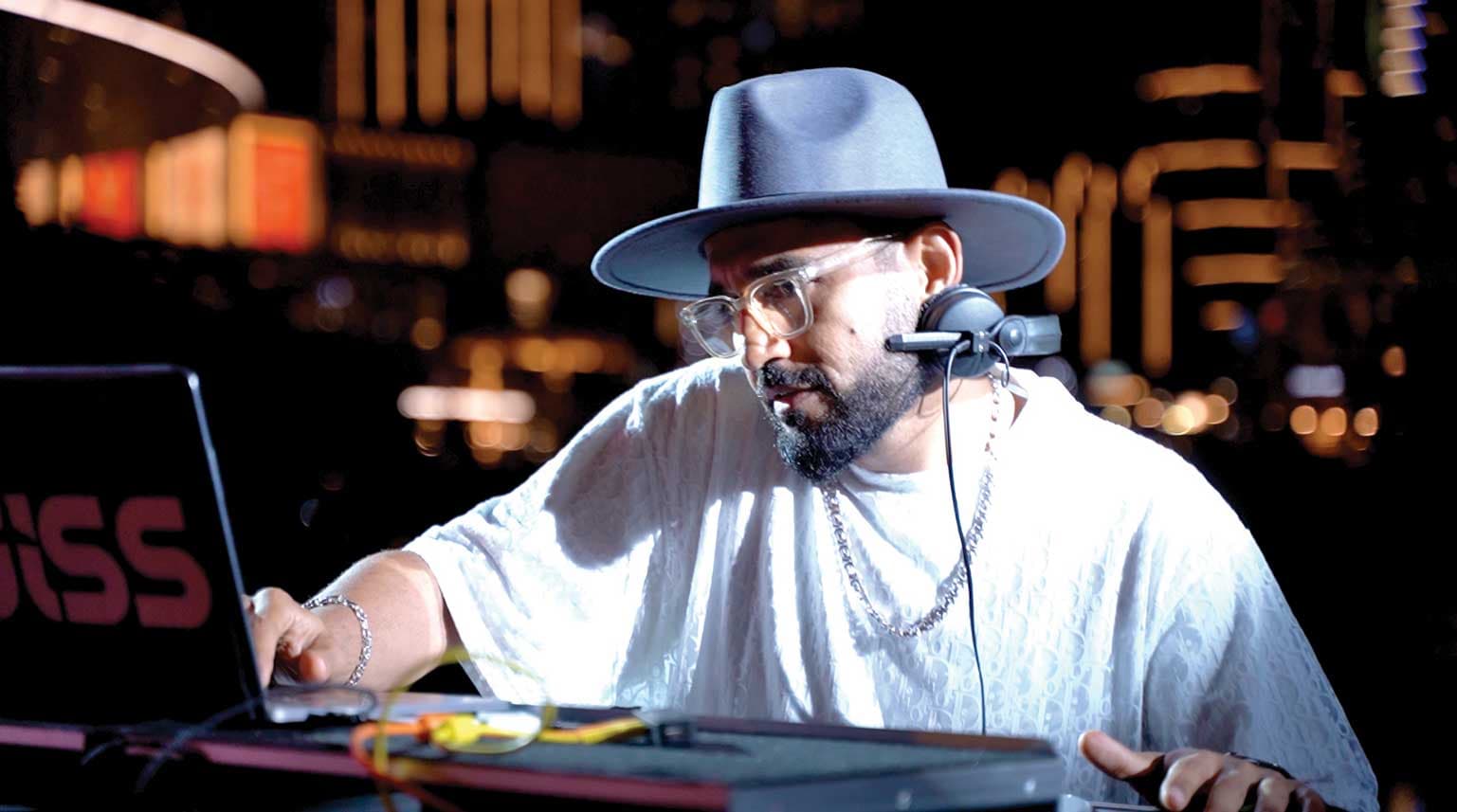 DJ wearing a wide-brim hat and headphones works on a laptop at a nighttime event