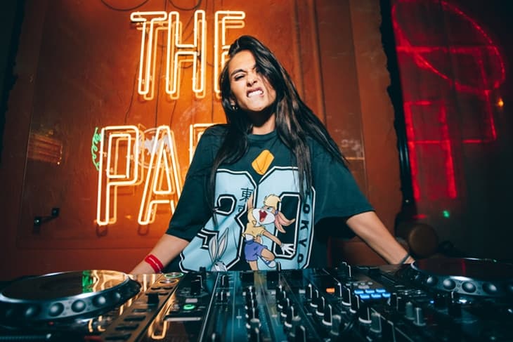 Woman DJ smiling behind a mixing console in a neon-lit club with a sign reading THE PA