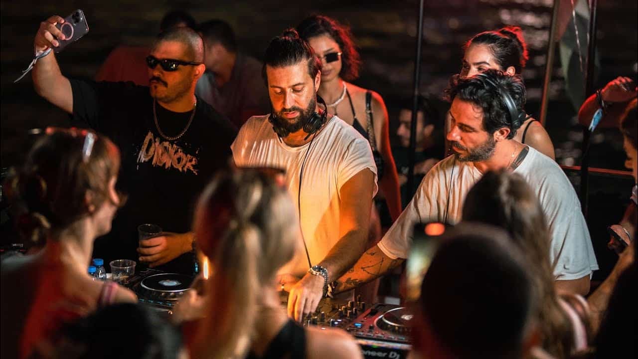 Three DJs perform at a crowded nightclub, with people filming them on their phones