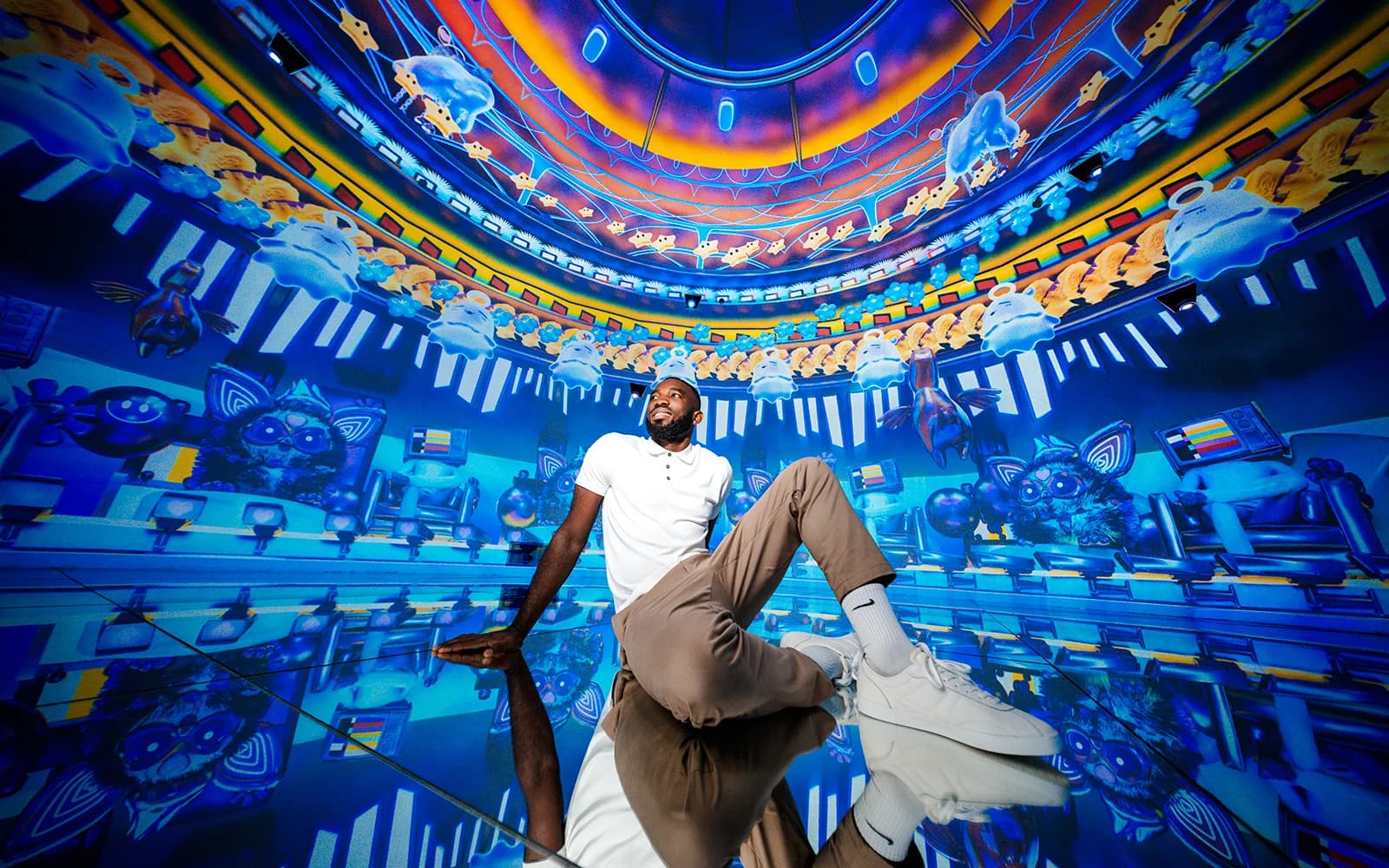 Man sitting on a reflective floor in a vivid blue and orange digital art installation with cartoon-like floating figures