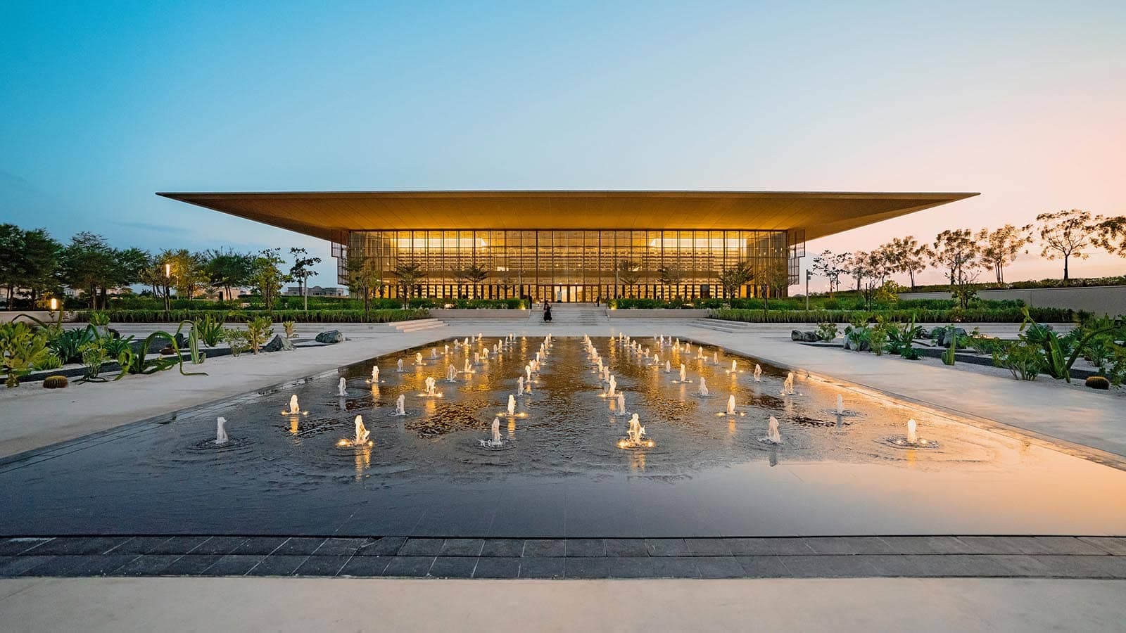 Modern glass building with a sweeping roof, reflected in a fountain plaza at dusk
