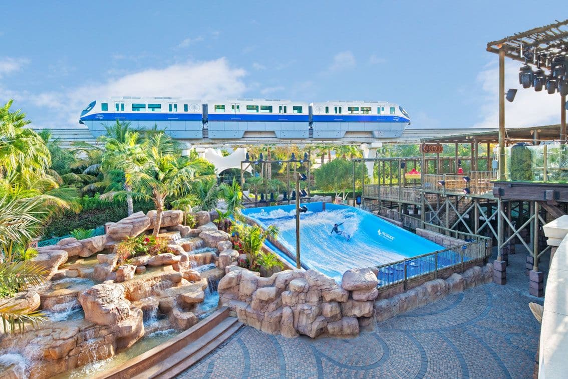 Elevated monorail passes over a tropical water park with a FlowRider surf simulator and rock waterfall