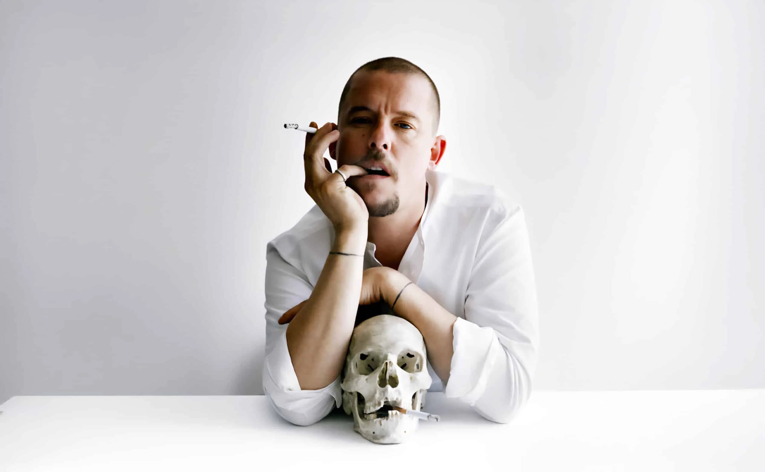 A person in a white shirt leans on a table holding a cigarette, with a skull positioned in front of them also holding a cigarette.
