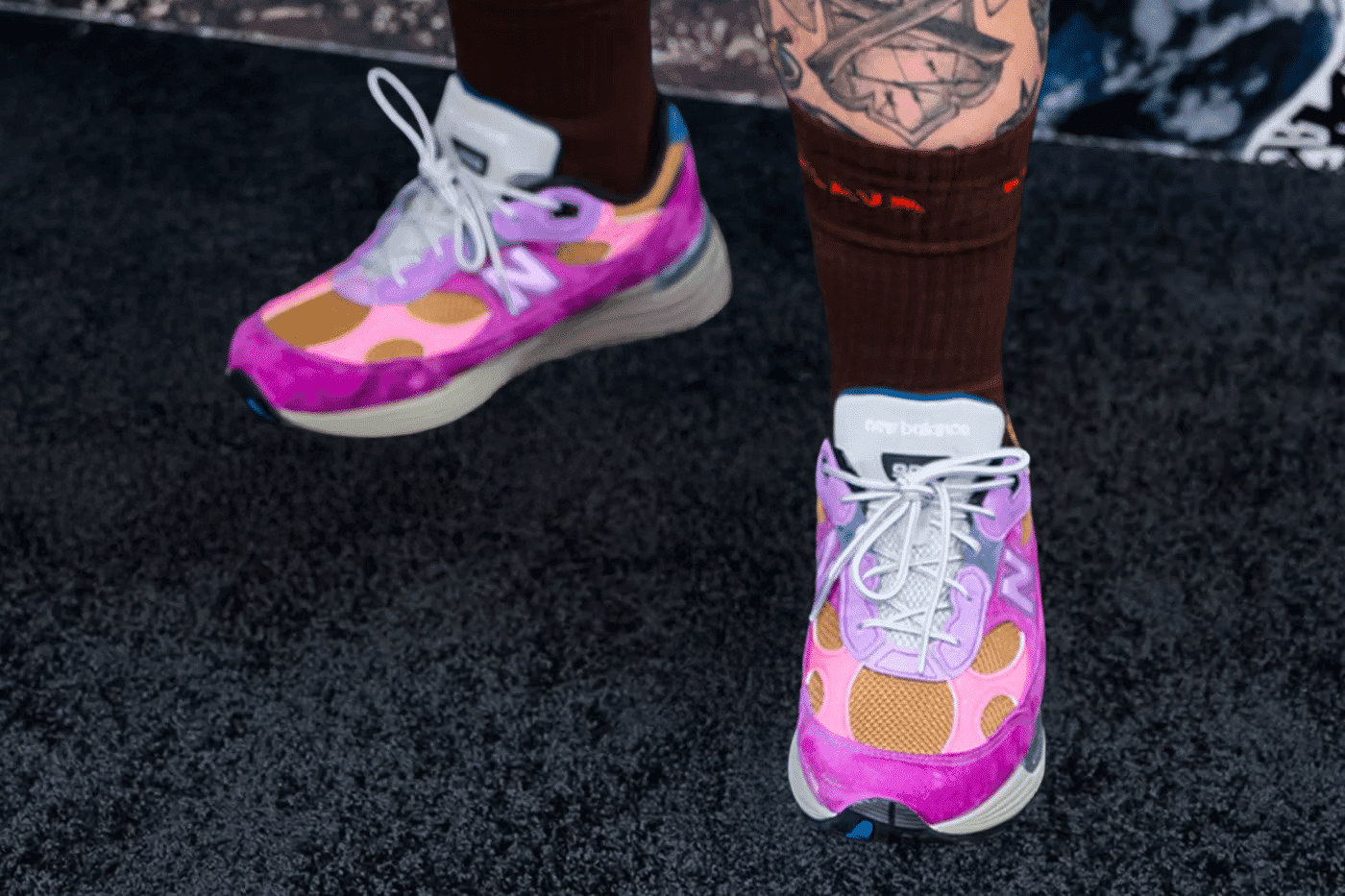 A pair of colorful sneakers with a pink, purple, and orange design, worn with brown socks and visible tattoos on the legs.